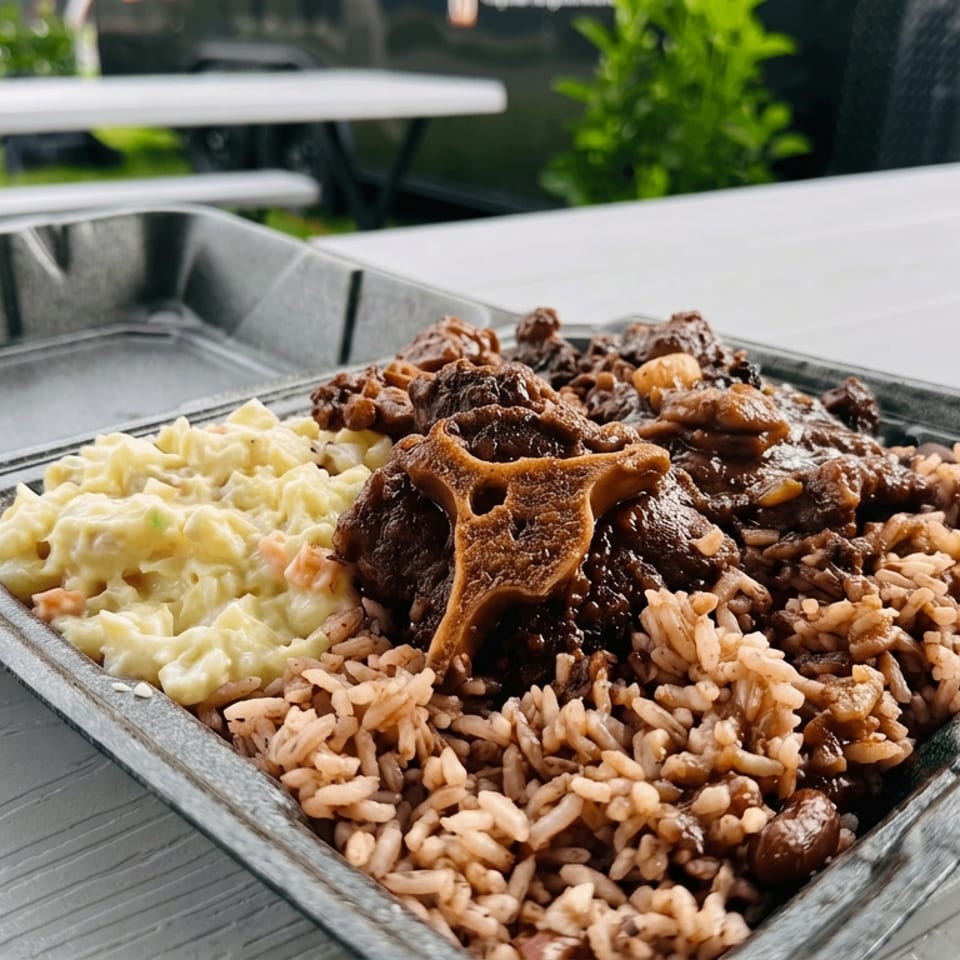 Takeout tray of braised oxtail with rice and peas, rice and gravy, and creamy macaroni salad on an outdoor picnic table.