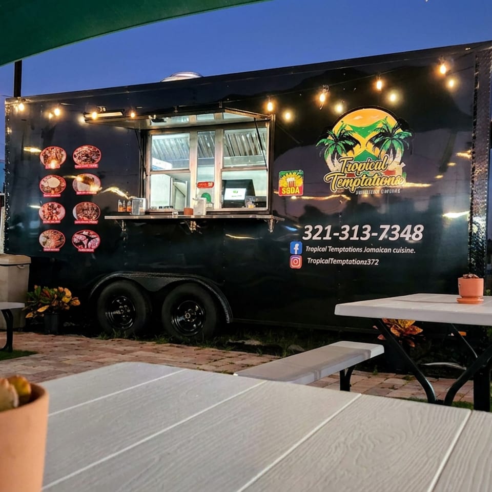 Tropical Temptations Jamaican cuisine food truck with illuminated string lights, picnic tables, and outdoor dining at dusk.