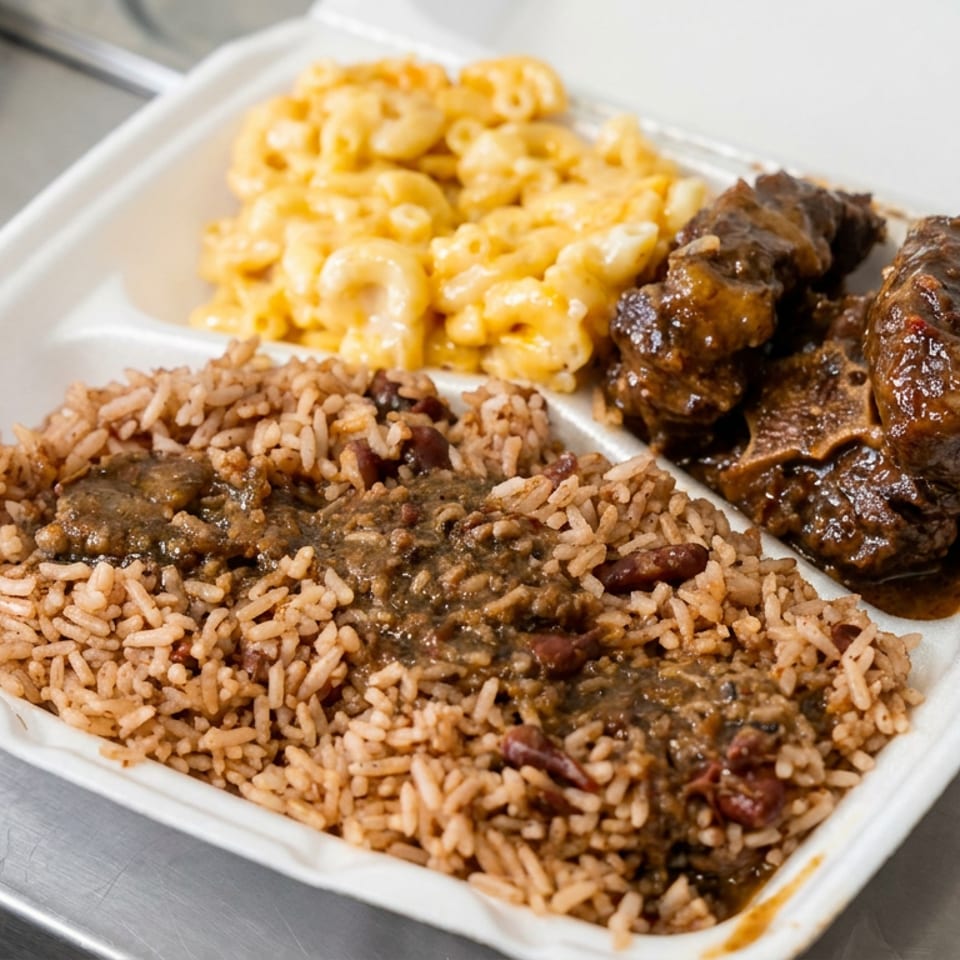 Takeout tray filled with seasoned rice and gravy, glazed meat, and creamy macaroni and cheese on a metal counter.